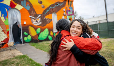 Two student artists, an older man and a younger woman, hug in front of a colorful wall.