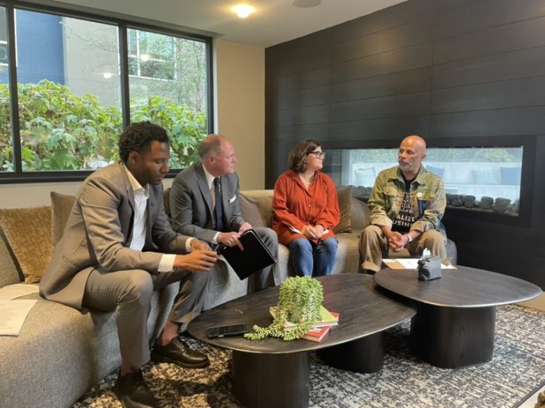 Four people sit on a couch and chairs in a modern room, engaged in conversation. Papers and a small plant are on the coffee table in front of them.