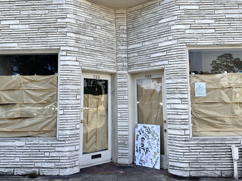 Two adjacent white brick storefronts with paper-covered windows; one door has a handwritten sign resting at its base, and a notice is posted on a window.
