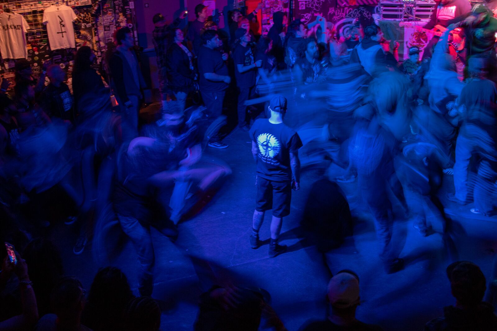 A man stands still in a mosh pit while people move in circles around him, their bodies blurry