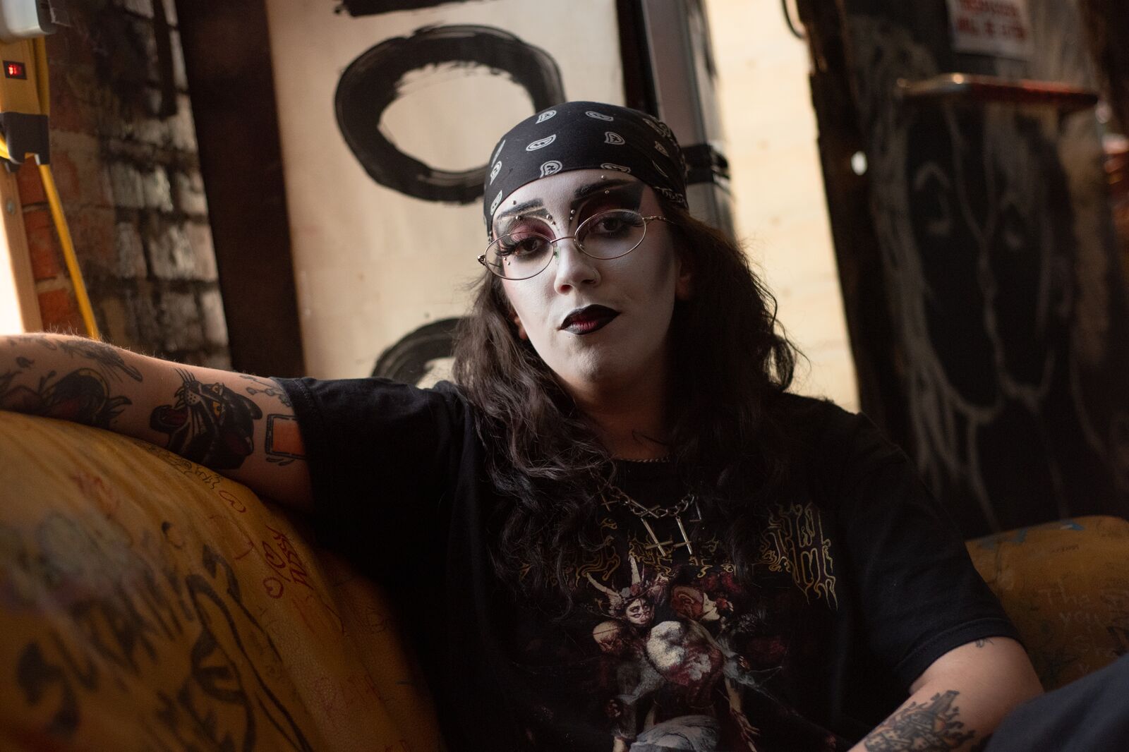A person wearing a black bandana and white makeup sits on a couch next to a window