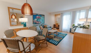 A modern hotel room suite with a round white table and four woven chairs, a pale blue couch with decorative pillows, large windows with sheer curtains, and a vibrant wall art piece.