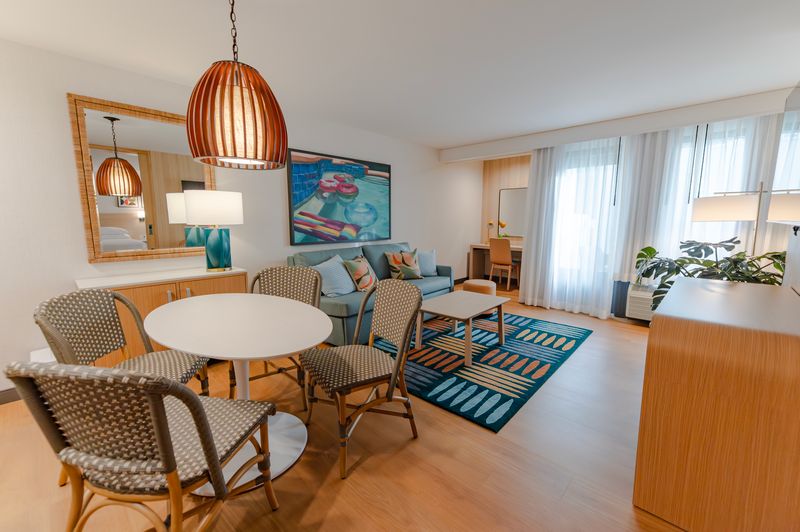 A modern hotel room suite with a round white table and four woven chairs, a pale blue couch with decorative pillows, large windows with sheer curtains, and a vibrant wall art piece.