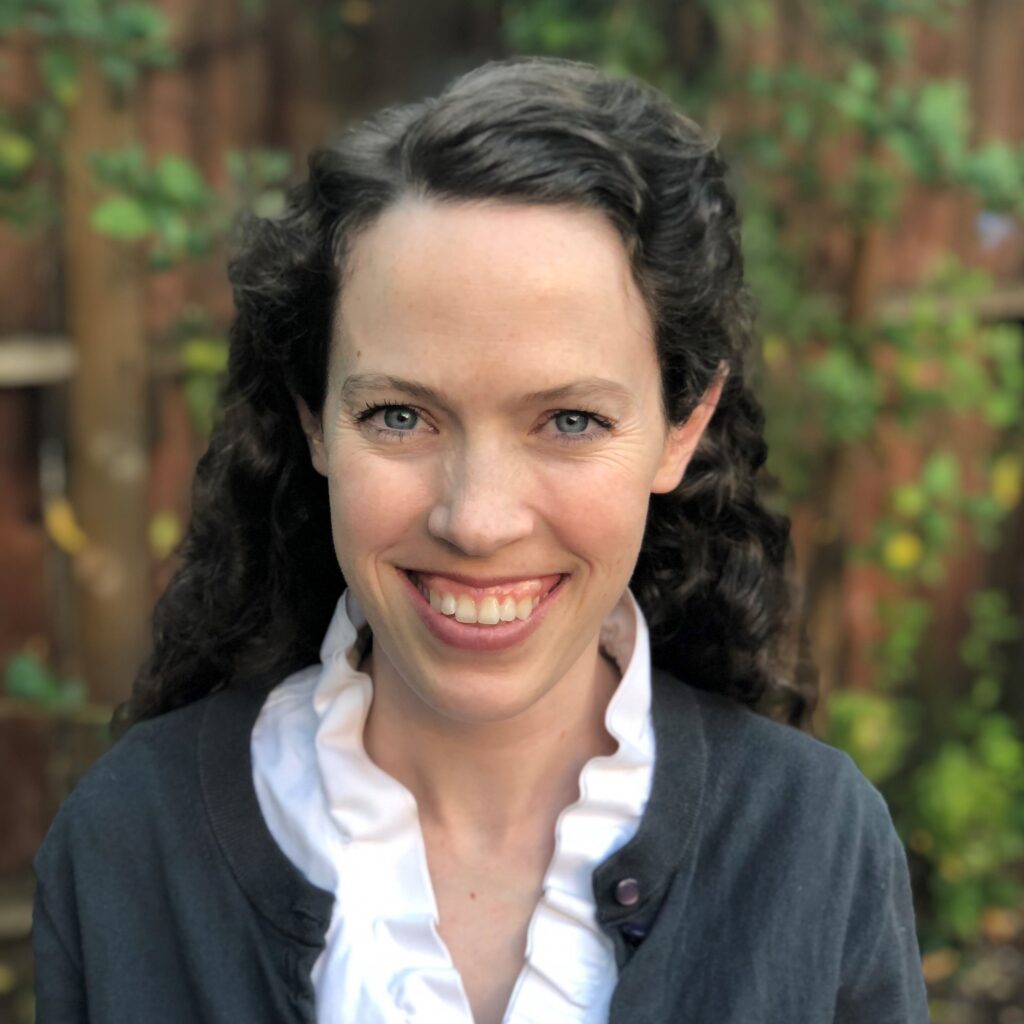 A close-up headshot portrait of a smiling woman with shoulder-length, dark curly hair and blue eyes looking directly at the camera. She wears a charcoal gray button-up cardigan sweater over a white collared shirt with a ruffled neckline. The background consists of softly blurred outdoor green foliage and a dark wooden fence.