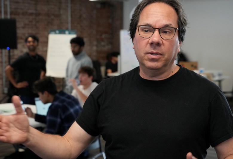 A person in a black shirt and glasses gestures while speaking in a workspace with others working in the background.