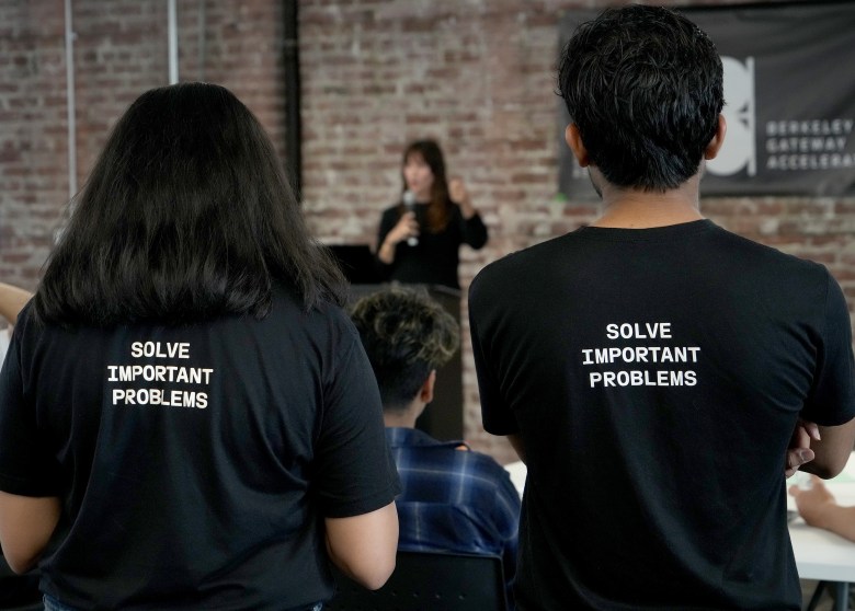 Two individuals wearing black shirts with "SOLVE IMPORTANT PROBLEMS" printed on the back stand facing a speaker at an event.