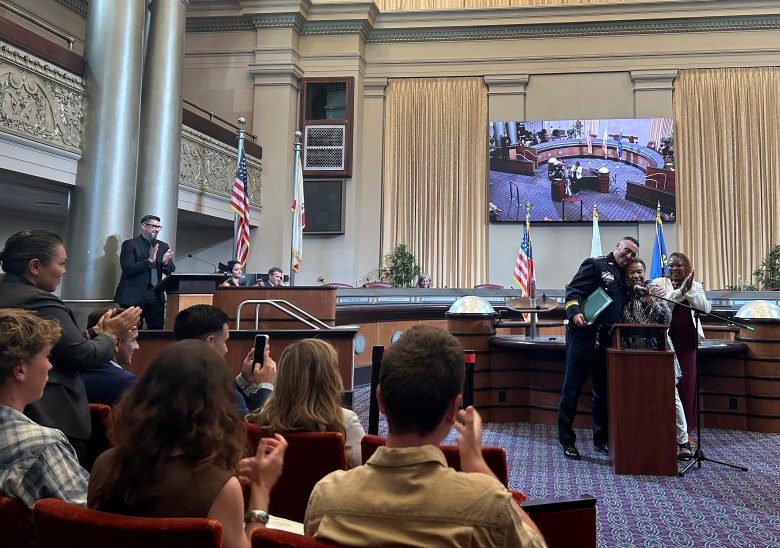 A uniformed officer and two others stand at a podium in a formal hall, with an audience applauding and a large screen displaying the same scene.