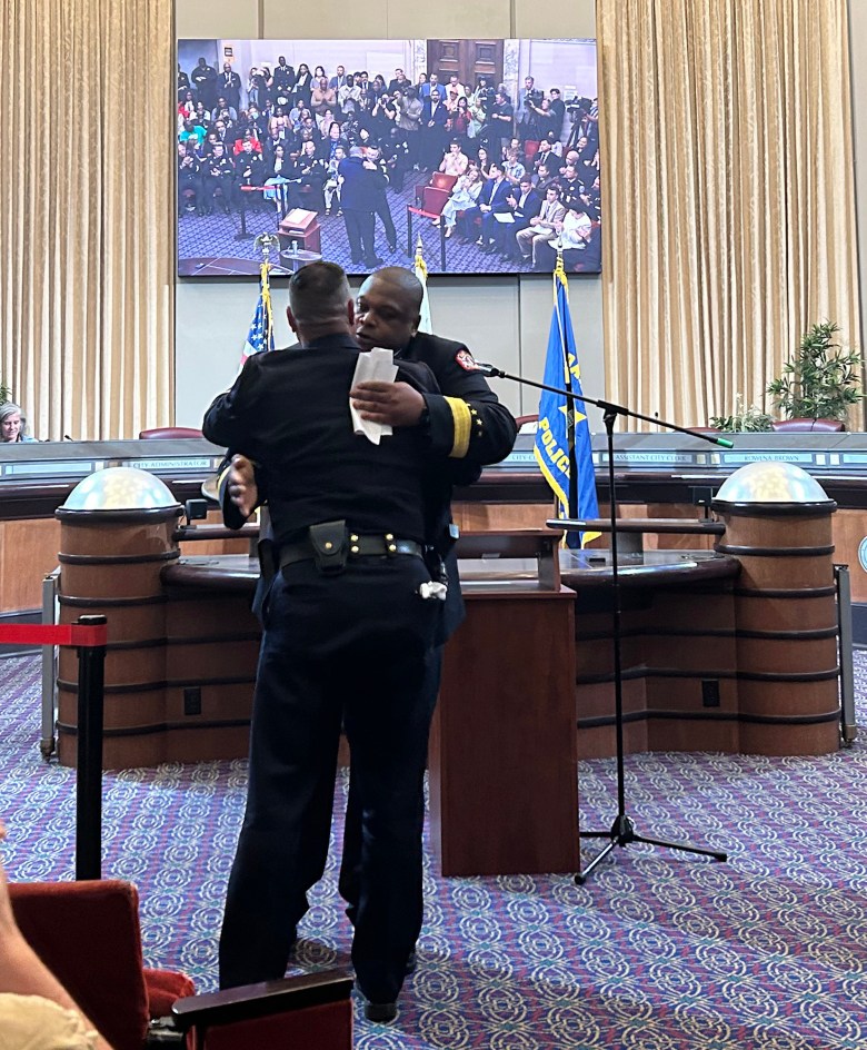 Two uniformed officers embrace warmly in a formal hall, with a large screen displaying an audience behind them.