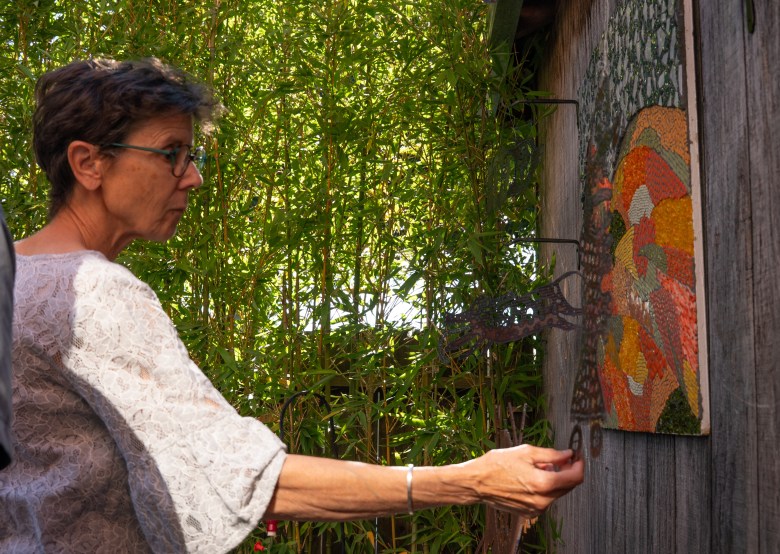 An individual wearing glasses and a lace top carefully places a small metal piece onto a colorful mosaic artwork mounted on a wooden wall, with tall green bamboo in the background.