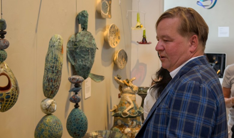 A person in a plaid jacket observes a wall display of colorful, handcrafted ceramic sculptures and decorative objects.