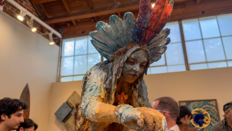 A textured, colorful sculpture of a figure with an elaborate headdress stands in an art gallery, surrounded by visitors.