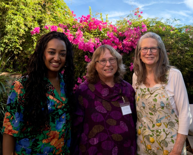 Three individuals pose together outdoors in front of vibrant pink flowers, each dressed in colorful, patterned attire.
