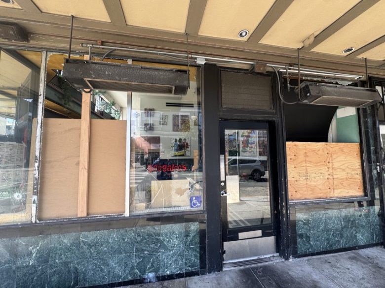 Storefront with boarded-up windows and door, green marble lower facade, a “50 gallons” sign inside, and a visible wheelchair accessibility sticker on the glass door.