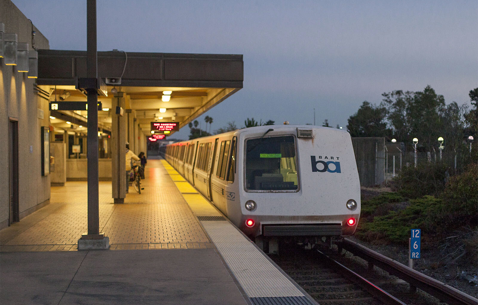 A new BART train sits on a track at night. To its left, a few people standn on the platform.