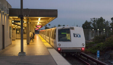 A new BART train sits on a track at night. To its left, a few people standn on the platform.