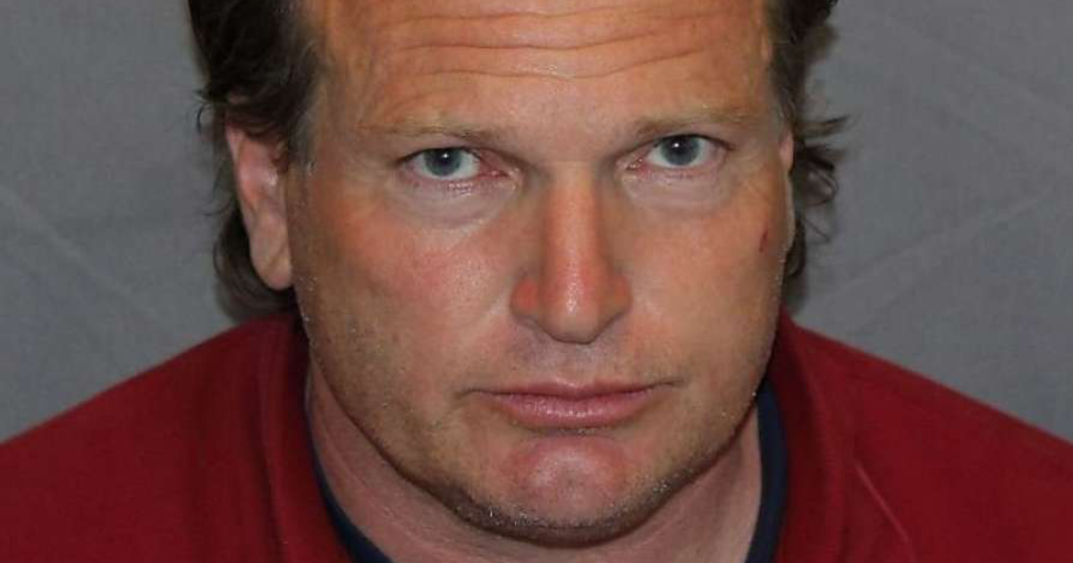 A middle-aged man with light blue eyes, receding brown hair, and a serious expression wears a red collared shirt over a dark blue undershirt against a plain gray background.