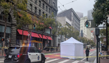 A police car blocks a city street with a white tent and yellow caution tape surrounding an area on a crosswalk, with officers and bystanders nearby.