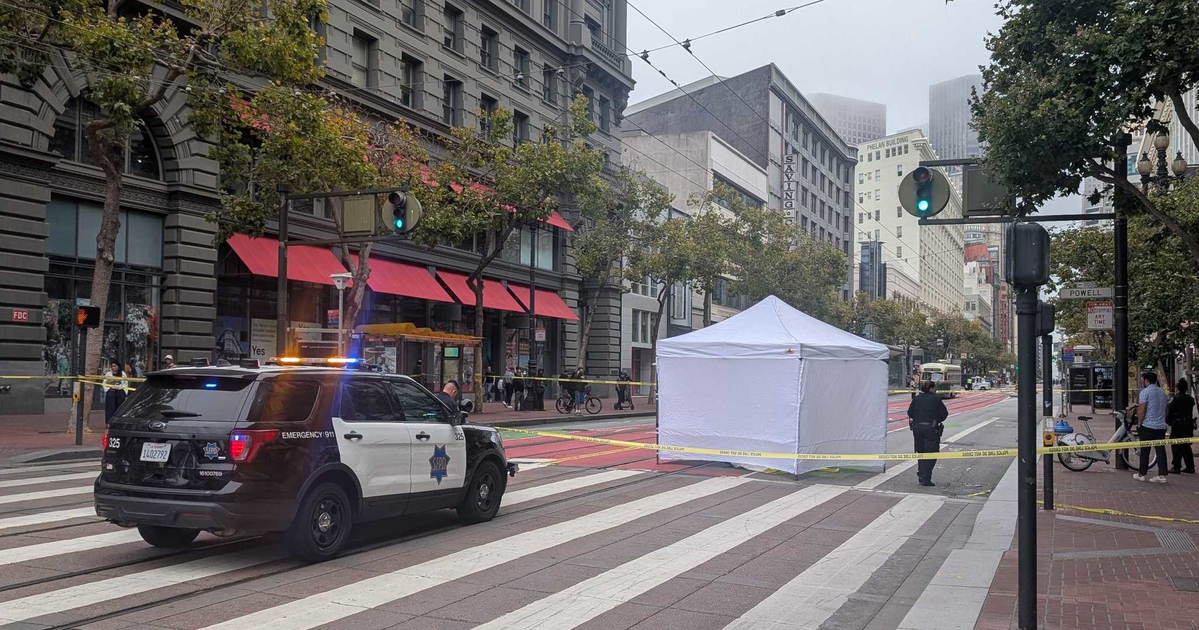 A police car blocks a city street with a white tent and yellow caution tape surrounding an area on a crosswalk, with officers and bystanders nearby.