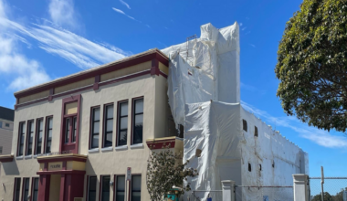 A two-story building with maroon trim and multiple windows stands next to a large structure covered in white plastic sheeting, with cars parked along the street.