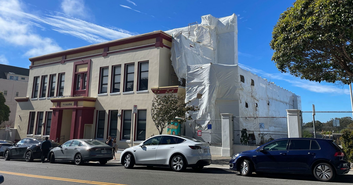 A two-story building with maroon trim and multiple windows stands next to a large structure covered in white plastic sheeting, with cars parked along the street.