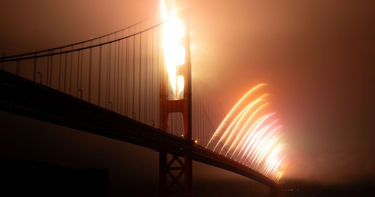 A bridge is illuminated by a series of bright, arcing fireworks that light up the night sky and reflect on the water below.
