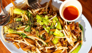 Stir-fried noodles with bean sprouts, green onions, and bok choy served on a white plate with a small cup of red chili sauce.