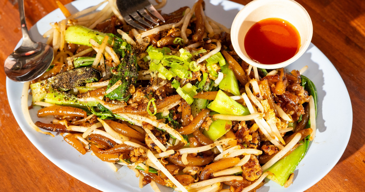 Stir-fried noodles with bean sprouts, green onions, and bok choy served on a white plate with a small cup of red chili sauce.