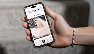 A hand holds a smartphone displaying an app called "Solve SF," showing a photo of a cleaned sidewalk under "Hall of Fame." A beaded bracelet is on the wrist.