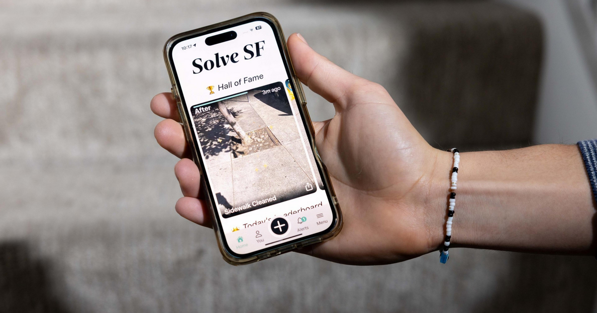 A hand holds a smartphone displaying an app called "Solve SF," showing a photo of a cleaned sidewalk under "Hall of Fame." A beaded bracelet is on the wrist.