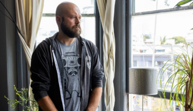 A bald man with a beard, wearing a black hoodie and jeans, sits on a couch with his hands clasped, looking thoughtfully to the side.