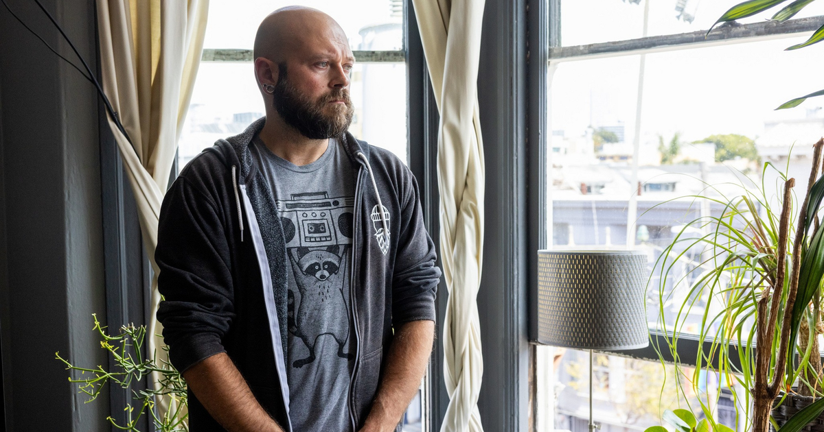 A bald man with a beard, wearing a black hoodie and jeans, sits on a couch with his hands clasped, looking thoughtfully to the side.