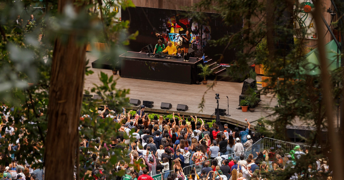 A music event is happening outdoors with a DJ on stage and a large crowd watching and raising their hands, surrounded by trees and greenery.