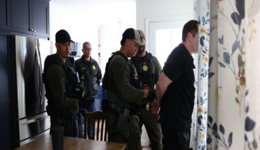 A man is handcuffed and held by three armed officers in tactical gear inside a kitchen, while a fourth officer stands nearby observing.