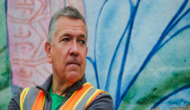 A middle-aged man in a neon orange safety vest and gray jacket stands with arms crossed in front of a colorful mural.