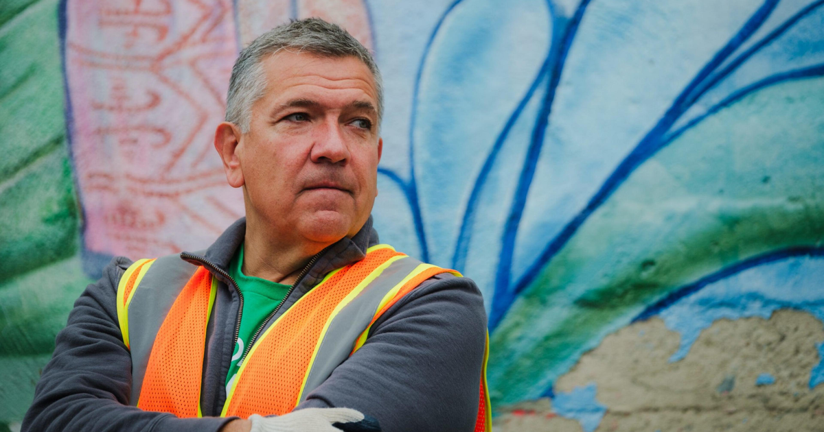 A middle-aged man in a neon orange safety vest and gray jacket stands with arms crossed in front of a colorful mural.