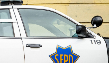 A white police car with the blue and gold emblem of "SFPD" on the side, marked with the number 119, parked against a yellow wall.
