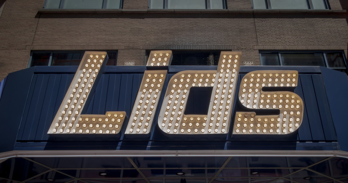 Large marquee lights spell out the word "Lids" on a dark blue sign mounted on a brick building.