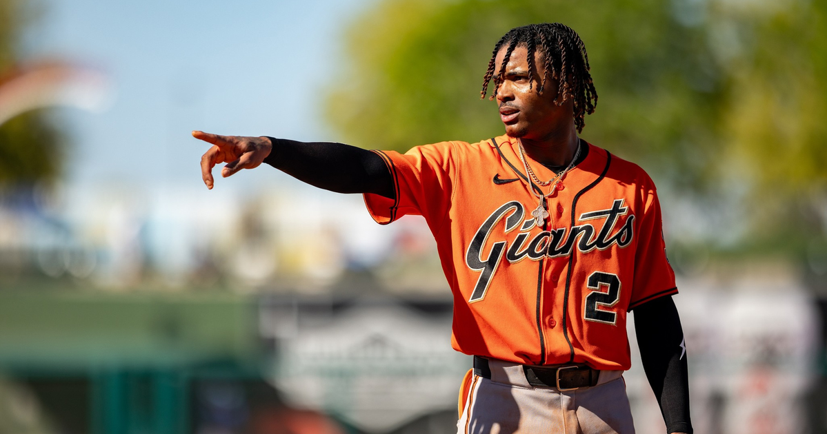 A baseball player in an orange "Giants" jersey with number 2 is pointing to his left on a sunny field.