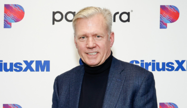 A middle-aged man with light hair, wearing a dark blazer and black turtleneck, stands smiling in front of a backdrop with Pandora and SiriusXM logos.