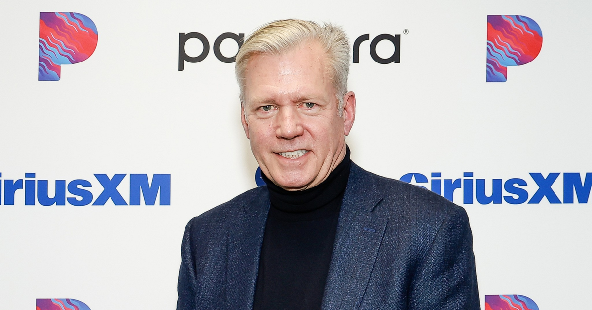 A middle-aged man with light hair, wearing a dark blazer and black turtleneck, stands smiling in front of a backdrop with Pandora and SiriusXM logos.