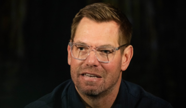 A man with short brown hair and clear glasses speaks, wearing a dark shirt against a dark, indistinct background.
