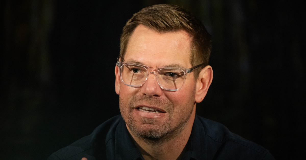 A man with short brown hair and clear glasses speaks, wearing a dark shirt against a dark, indistinct background.