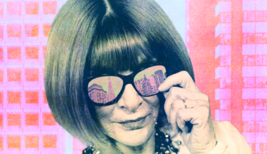 A woman with a bob haircut adjusts her large black sunglasses, reflecting city buildings, while wearing a chunky necklace against a pink cityscape background.