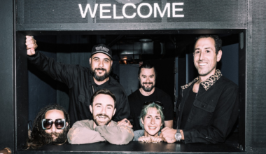 Six people stand casually on a sidewalk outside a building with a vertical "Folsom Street Laundry" sign hanging above them.