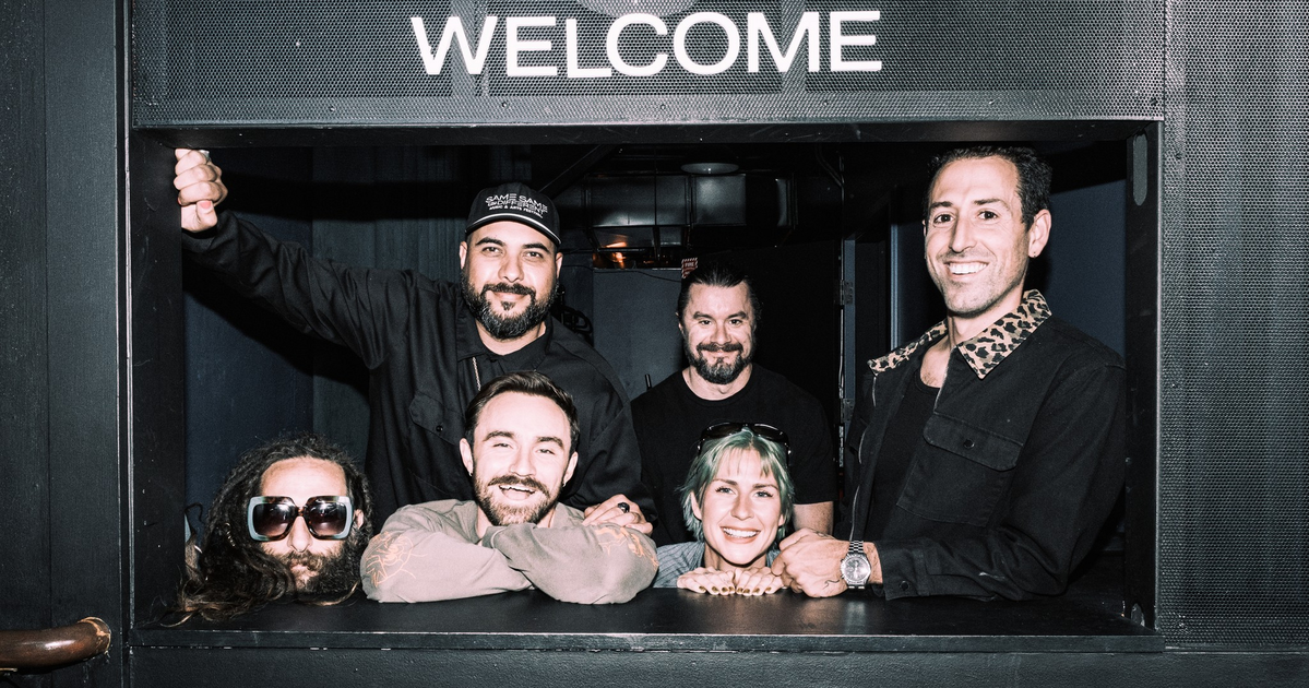 Six people stand casually on a sidewalk outside a building with a vertical "Folsom Street Laundry" sign hanging above them.