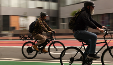 Two men wearing helmets ride bicycles on a green bike lane in an urban area with blurred buildings in the background.