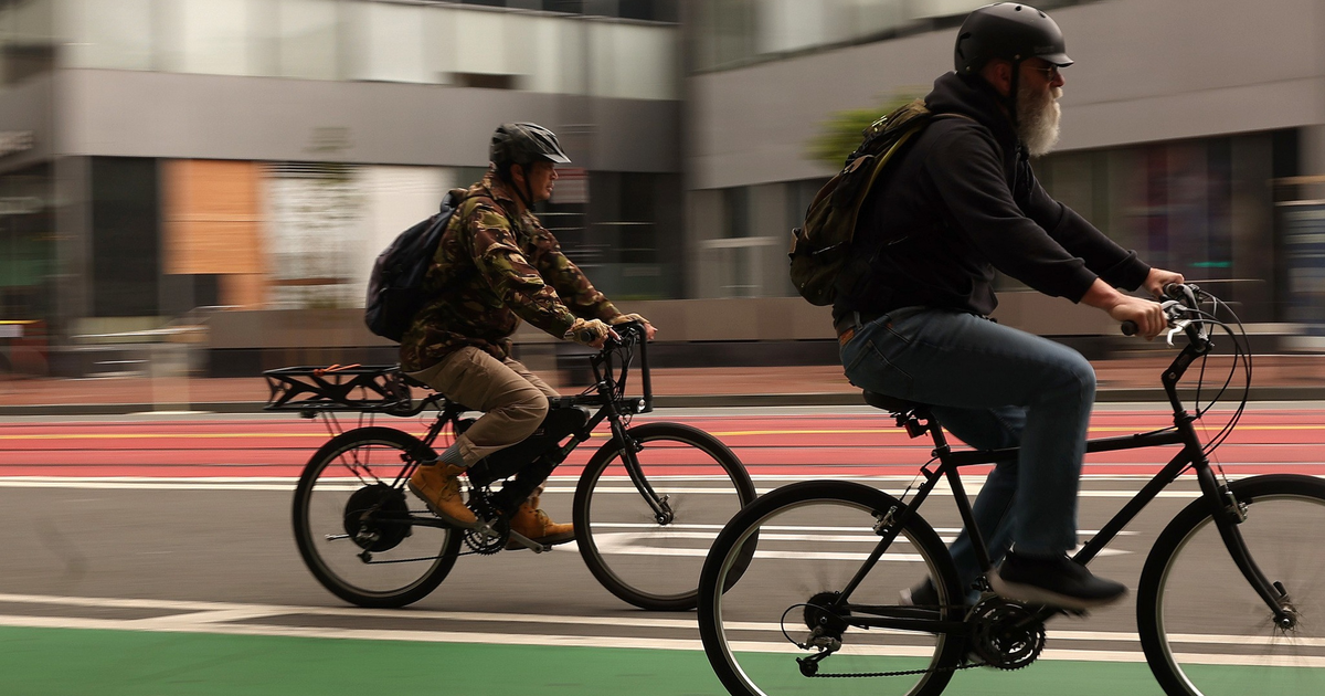 Two men wearing helmets ride bicycles on a green bike lane in an urban area with blurred buildings in the background.