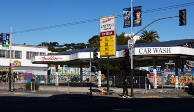 A closed car wash with graffiti-covered walls surrounds the structure, a speed limit sign, and a pedestrian walking by on a sunny day.