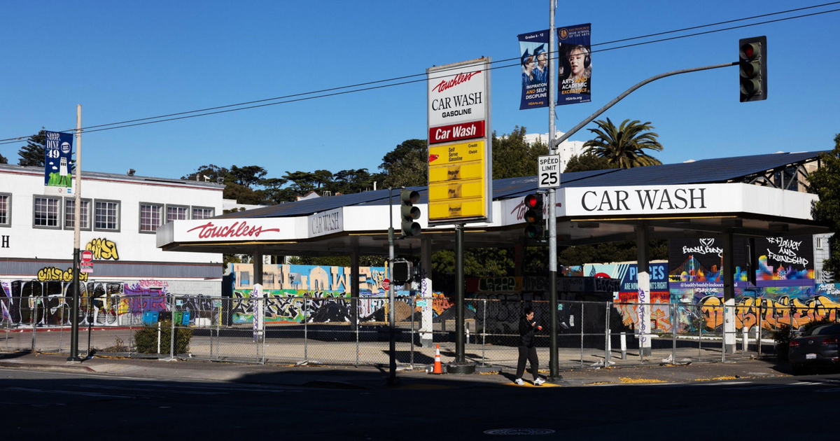 A closed car wash with graffiti-covered walls surrounds the structure, a speed limit sign, and a pedestrian walking by on a sunny day.