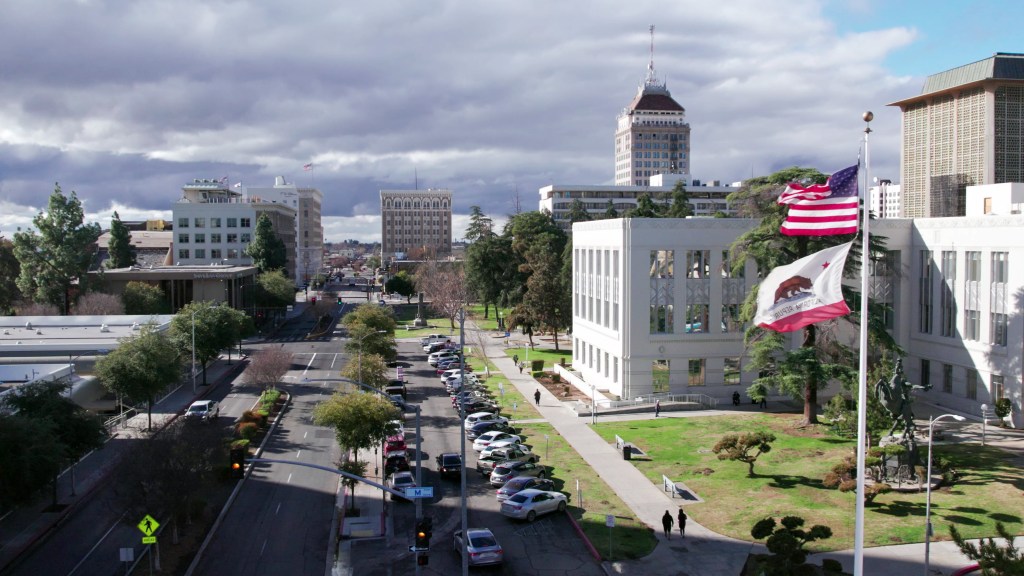 An aerial view of Courthouse Park in downtown Fresno, California, with the American and Californian flags flying, surrounded by city buildings and parked cars.